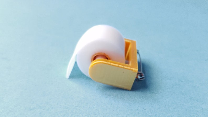 May include: A miniature gold-colored toilet paper holder with a white roll of paper.