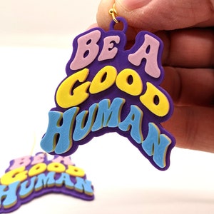 May include: Purple, blue, yellow, and pink acrylic earrings with the words "Be a Good Human" in a retro font.