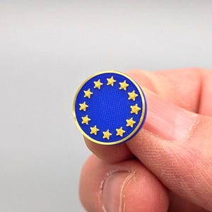 Pode incluir: Um alfinete circular azul com contorno dourado e 12 estrelas douradas dispostas em círculo.