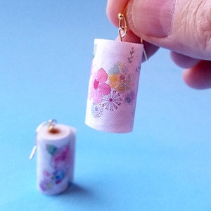 May include: A pair of dangle earrings featuring miniature paper towel rolls with a floral print. The paper towel rolls are white with pink, yellow, and blue flowers.