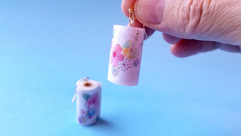 May include: A pair of dangle earrings featuring miniature paper towel rolls with a floral print. The paper towel rolls are white with pink, yellow, and blue flowers.
