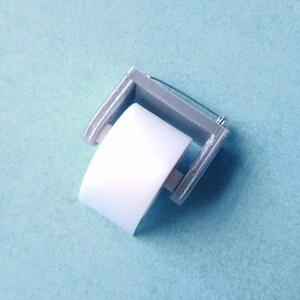 May include: A miniature silver-colored toilet paper holder with a white paper roll. The holder is designed to look like a traditional toilet paper holder.