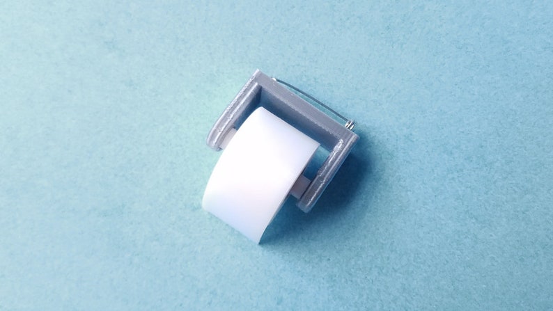 May include: A miniature silver-colored toilet paper holder with a white paper roll. The holder is designed to look like a traditional toilet paper holder.