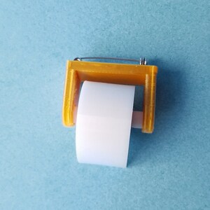 May include: A small, yellow plastic dispenser with a white tape roll. The dispenser has a metal pin on top.
