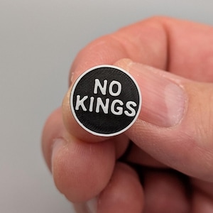 Puede incluir: Un pequeño botón redondo en blanco y negro con las palabras "NO KINGS" en letras blancas. El botón se sostiene entre los dedos. El botón tiene una superficie texturizada y un fondo neutro.