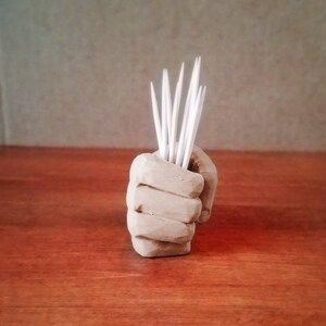 Pu&ograve; includere: Una scultura di mano in ceramica marrone che tiene un set di stuzzicadenti bianchi.