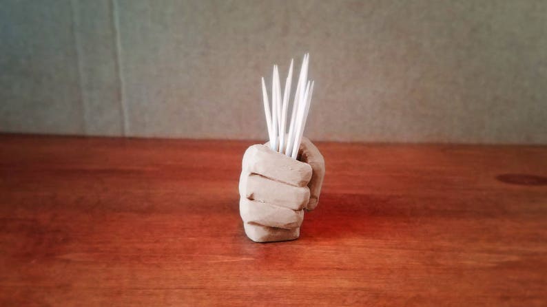 Pu&ograve; includere: Una scultura di mano in ceramica marrone che tiene un set di stuzzicadenti bianchi.