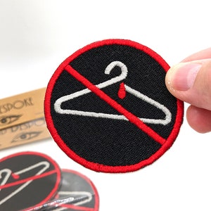 May include: A red and black embroidered patch with a white hanger and a red drop of blood. The hanger is crossed out with a red line. The text "Woke and Despoke" is visible in the background.