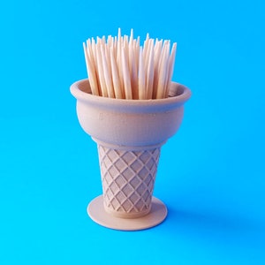 Op de afbeelding: Een bruine, keramische tandenstokerhouder in de vorm van een ijshoorn met houten tandenstokers erin. De houder staat op een blauwe achtergrond.