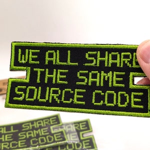 May include: A black and green embroidered patch with the text "WE ALL SHARE THE SAME SOURCE CODE" in a pixelated font.