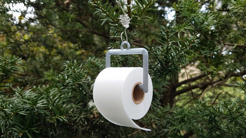 May include: A white toilet paper roll ornament hanging from a tree branch. The ornament is made of a gray 3D printed holder with a metal hook.
