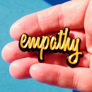 May include: A black acrylic pin with the word "empathy" written in gold lettering.