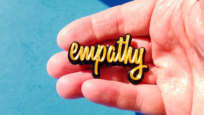 May include: A black acrylic pin with the word "empathy" written in gold lettering.
