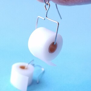 May include: A pair of silver earrings shaped like toilet paper rolls. The rolls are white and have a brown cardboard center.