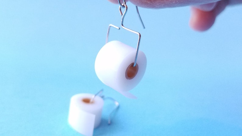 May include: A pair of silver earrings shaped like toilet paper rolls. The rolls are white and have a brown cardboard center.