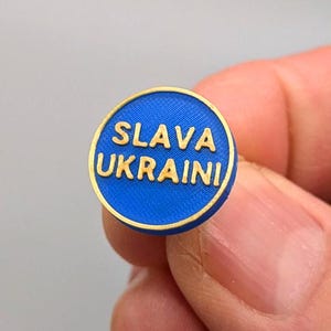 Pode incluir: Um alfinete redondo azul com letras douradas que diz "SLAVA UKRAINI".