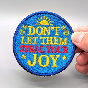 May include: A circular embroidered patch with a blue background and a dark blue border. The text DON'T LET THEM in yellow, STEAL YOUR in red, and JOY in yellow is centered. A yellow sun icon is at the top, with yellow swirls and stars.