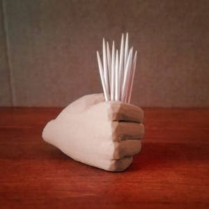 Pu&ograve; includere: Una scultura di mano in ceramica beige con un gruppo di stuzzicadenti bianchi che sporgono dalla parte superiore.