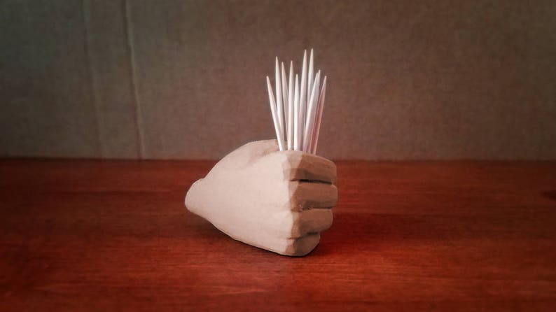 Pu&ograve; includere: Una scultura di mano in ceramica beige con un gruppo di stuzzicadenti bianchi che sporgono dalla parte superiore.