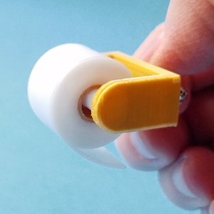 May include: A small, yellow plastic dispenser holds a roll of clear plastic wrap. The dispenser has a handle and a small screw on the side.