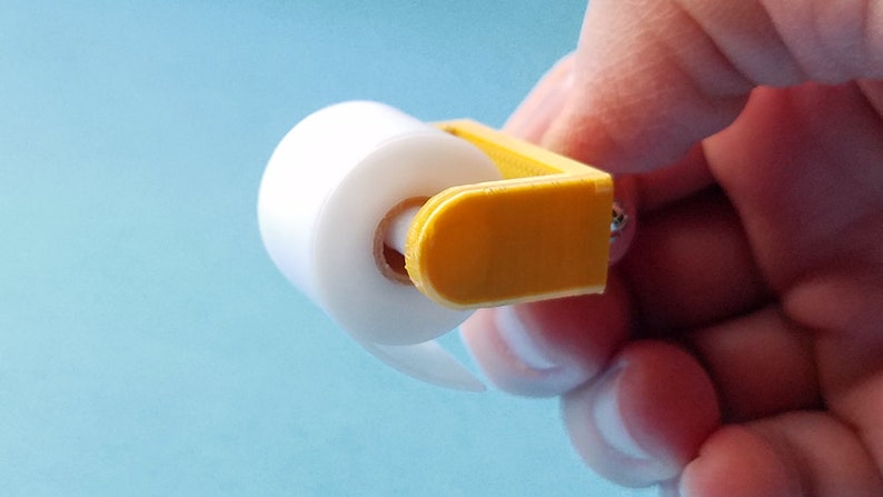 May include: A small, yellow plastic dispenser holds a roll of clear plastic wrap. The dispenser has a handle and a small screw on the side.