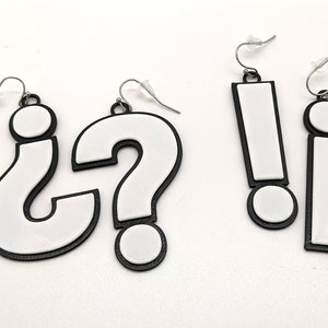 Puede incluir: Tres pares de pendientes con símbolos de signo de interrogación, signo de exclamación y la letra minúscula 'i'. Los pendientes son blancos con contornos negros y están hechos de plástico.