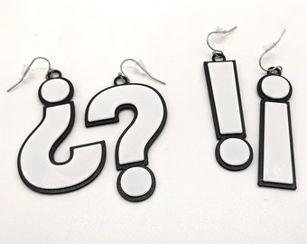 May include: Three pairs of earrings featuring question mark, exclamation point, and lowercase letter 'i' symbols. The earrings are white with black outlines and are made of plastic.