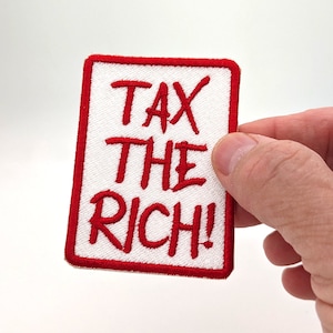 May include: Embroidered patch with the text "TAX THE RICH!" in red on a white background, framed by a red border. The patch is rectangular and held by a hand against a white backdrop.