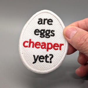 May include: White egg-shaped patch with black and red embroidered text that reads "are eggs cheaper yet?"