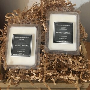 May include: Two rectangular wax melt containers labeled "Salted Caramel" from Mason Jar Candles & Co. are nestled in a wooden crate filled with brown paper shreds. Each container holds a white wax melt and includes the text "100% Soy Wax" and "2.8 oz".