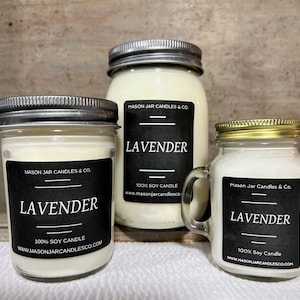 Three candles, a 4oz, 8oz and 16oz jar are on a soft white fabric. A light brown board is behind candles. Candle scent is Lavender. Candle label is black, with white writing.