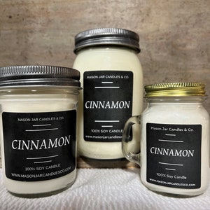 May include: Three mason jar candles with silver and gold lids. Each candle has a black label with the word "CINNAMON" in white. The labels also include "100% SOY CANDLE" and the website address. The jars are filled with a creamy wax.
