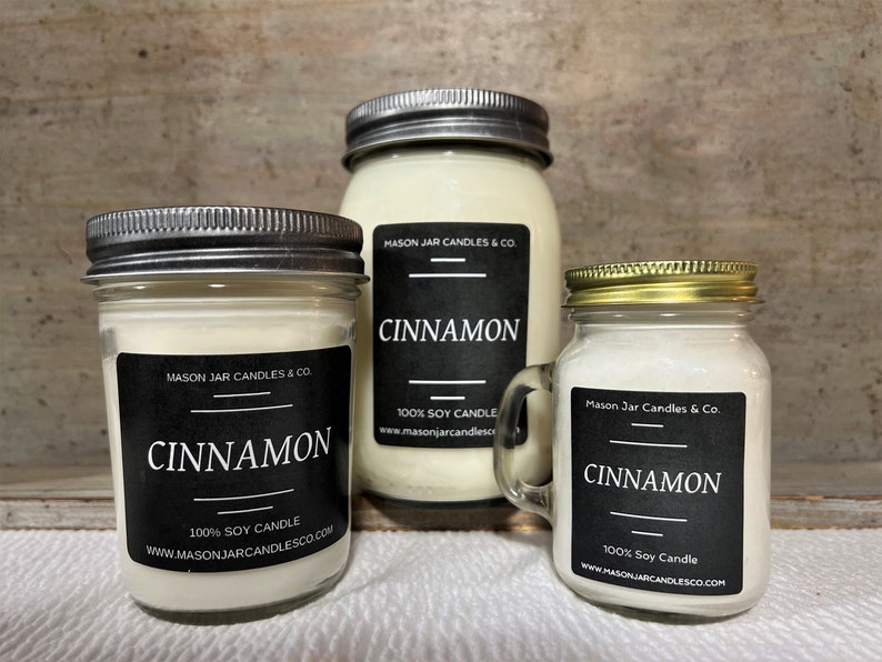 Three candles, a 4oz, 8oz and 16oz jar are on a soft white fabric. A light brown board is behind candles. Candle scent is Cinnamon. Candle label is black, with white writing.