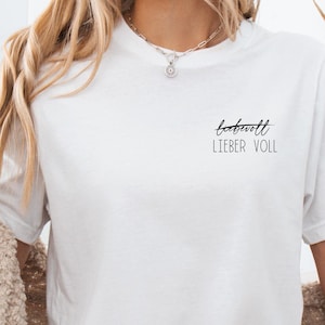 Könnte beinhalten: Ein weißes T-Shirt mit dem Aufdruck "bekannt lieber voll" auf der Vorderseite. Das Shirt wird von einer Person mit langen blonden Haaren getragen und zu Jeans-Shorts und einem cremefarbenen Cardigan kombiniert.