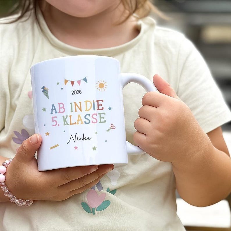 May include: White ceramic mug with a handle, decorated with colourful illustrations and text. The text includes "2026", "AB IN DIE 5. KLASSE", and a name. The mug is held by a person.