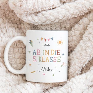 May include: White ceramic mug with a handle, decorated with colourful school-themed illustrations and text. The text includes "AB IN DIE 5. KLASSE" and "2026". Additional illustrations include a sun, stars, and scissors.
