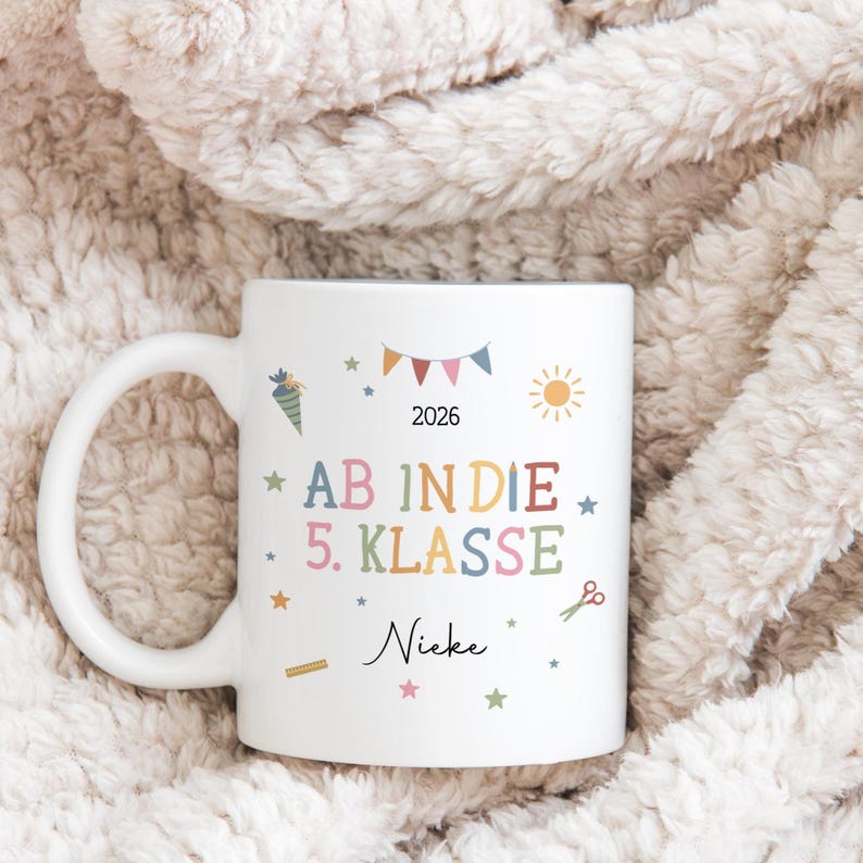 May include: White ceramic mug with a handle, decorated with colourful school-themed illustrations and text. The text includes "AB IN DIE 5. KLASSE" and "2026". Additional illustrations include a sun, stars, and scissors.