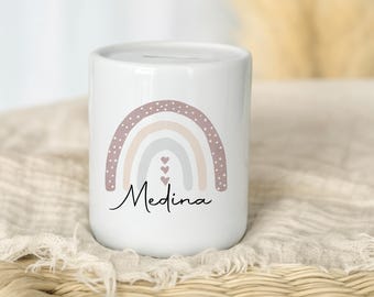 Könnte beinhalten: Weiße Keramik-Spardose mit einem Regenbogen-Design in Rosa-, Beige- und Grautönen. Das Wort "Medina" ist in Schreibschrift unter dem Regenbogen geschrieben. Die Spardose hat oben einen Schlitz zum Einwerfen von Münzen.