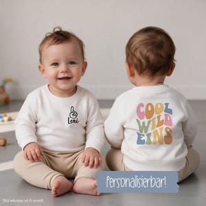 Könnte beinhalten: Ein cremefarbenes Langarm-Sweatshirt mit einer kleinen schwarzen Grafik eines nach oben zeigenden Fingers und dem Namen Leni auf der Vorderseite sowie dem Text COOL WILD EINS in bunten, wellenförmigen Retro-Buchstaben auf der Rückseite.