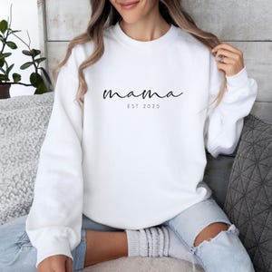 Könnte beinhalten: Weißes Sweatshirt mit Rundhalsausschnitt, auf dem das Wort "mama" in Schreibschrift und darunter "EST 2025" steht. Das Sweatshirt ist einfarbig weiß. Ein lässiges, bequemes Kleidungsstück.