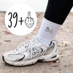 Könnte beinhalten: Weiße Sportsocken mit dem Text "39+" und einer Mittelfinger-Grafik. Die Socken werden mit schwarzen Leggings und einem silber-weißen Sportschuh getragen. Ein weißer Kreis mit dem gleichen Text und der gleichen Grafik befindet sich im Hintergrund.
