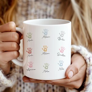 May include: White ceramic mug featuring colorful handprints and names. The handprints are in pastel shades, with names such as Noah, Milan, and Smilla. The mug is held by a person.