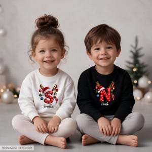 Puede incluir: Una sudadera blanca de manga larga con una letra S roja con astas de reno y adornos navideños, y una sudadera negra de manga larga con una letra N roja con astas de reno y adornos navideños, ambas con nombres impresos.