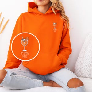 Peut inclure: Un sweat à capuche orange vif avec un petit motif de cocktail sur la poitrine et un grand motif circulaire sur le devant. Le motif circulaire porte l'inscription "IT'S SPRITZ O'CLOCK". Style décontracté.