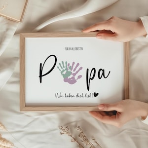 Könnte beinhalten: Ein gerahmter Druck mit dem Wort "Papa" in schwarzer Schrift, mit drei Handabdrücken in Grün, Lila und Lavendel. Der Text "Für den Allerbesten" und "Wir haben dich lieb!" sind ebenfalls abgebildet. Der Rahmen ist hellbraun.
