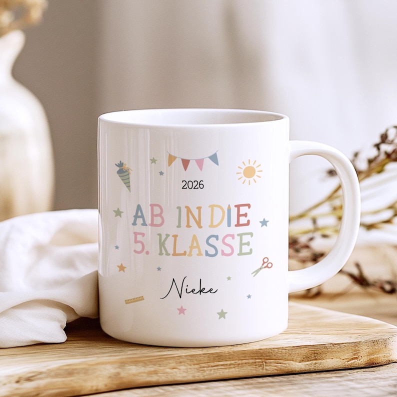 May include: White ceramic mug with the text "AB IN DIE 5. KLASSE" in colourful letters, the year "2026" and the name "Nieke". The mug is decorated with stars, a carrot, scissors, and a sun.