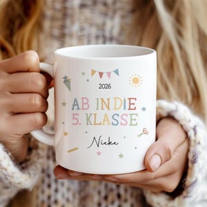 May include: White ceramic mug with colourful text and graphics. The mug features the words "AB IN DIE 5. KLASSE" and the year "2026". Additional graphics include stars, a carrot, a sun, and scissors. The name "Nieke" is also printed on the mug.