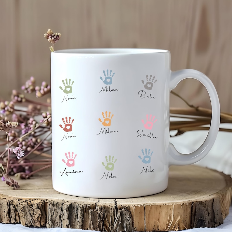 May include: White ceramic mug decorated with colourful handprints and names. The handprints are in shades of green, orange, pink, and blue. Each handprint has a name written below it in a cursive font. The mug has a curved handle.