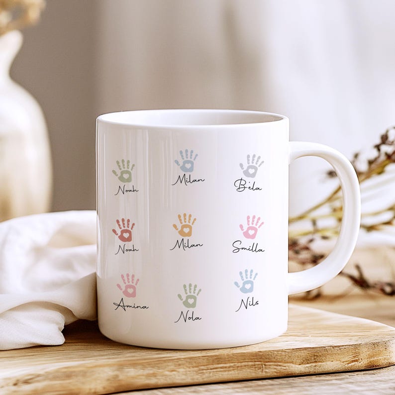 May include: White ceramic mug with pastel-coloured handprints and names such as Noah, Milan, and Smilla. The handprints are in shades of pink, blue, green, and orange. The mug has a curved handle and is on a wooden surface.