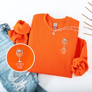 Pode incluir: Camisola laranja brilhante com um gráfico de cocktail e o texto "IT'S SPRITZ O'CLOCK". A camisola é combinada com jeans azuis e um colar. Um close-up mostra o design do cocktail.